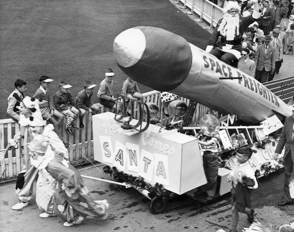 The Santa float in the James Smith Christmas parade, Wellington. The photograph shows Santa Claus sitting in a space rocket attended by young spacemen. Clowns are pulling the float, and boys are sitting on the fence watching. Beyond the float can be seen part of the crowd. Photographed by K E Niven Ltd between 1962 and 1968. http://mp.natlib.govt.nz/detail/?id=21736