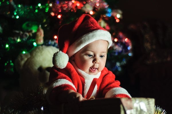 A baby in a Santa suit opens a present. Click the photo read about Christmas giving: what charities really want you to donate on the The Spinoff.